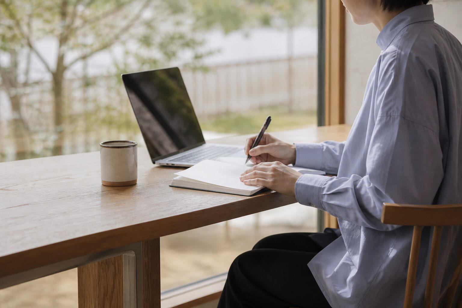 カフェの窓際でノートパソコンとノートを広げて学習する社会人